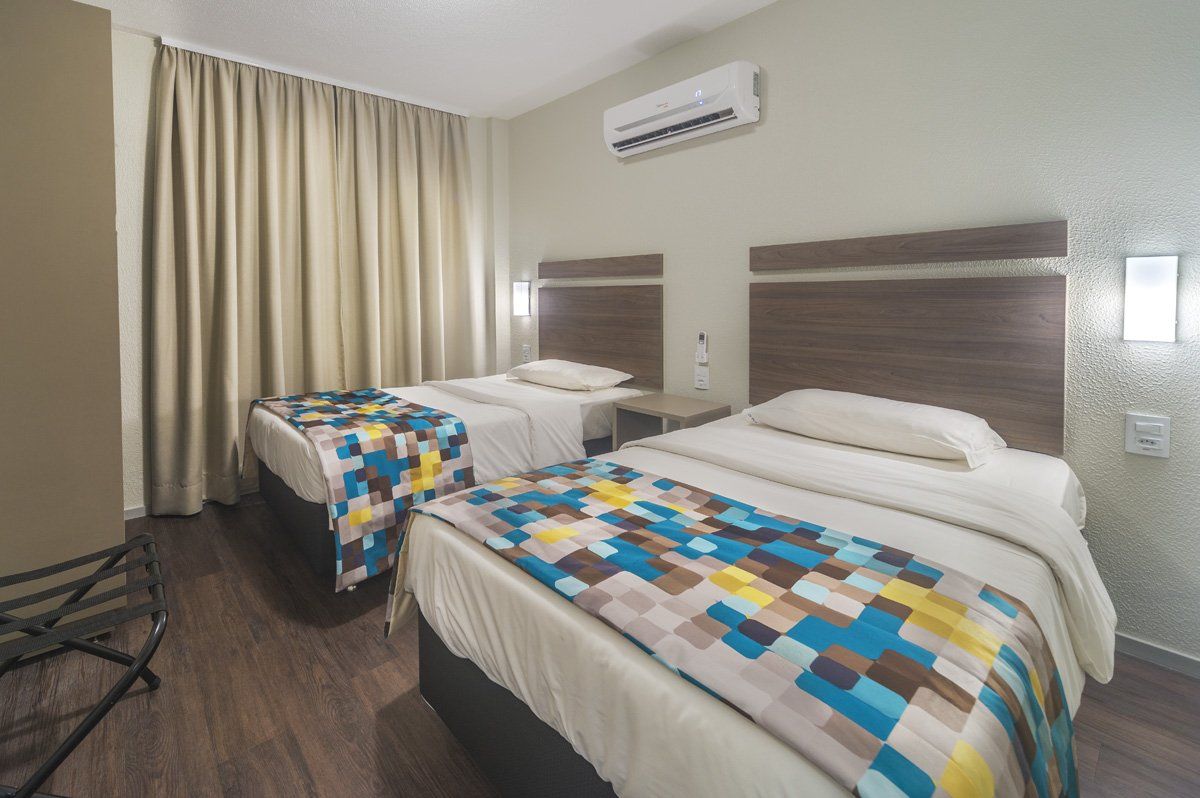 Quarto de hotel com duas camas de solteiro, cortinas bege e colchas coloridas de retalhos.