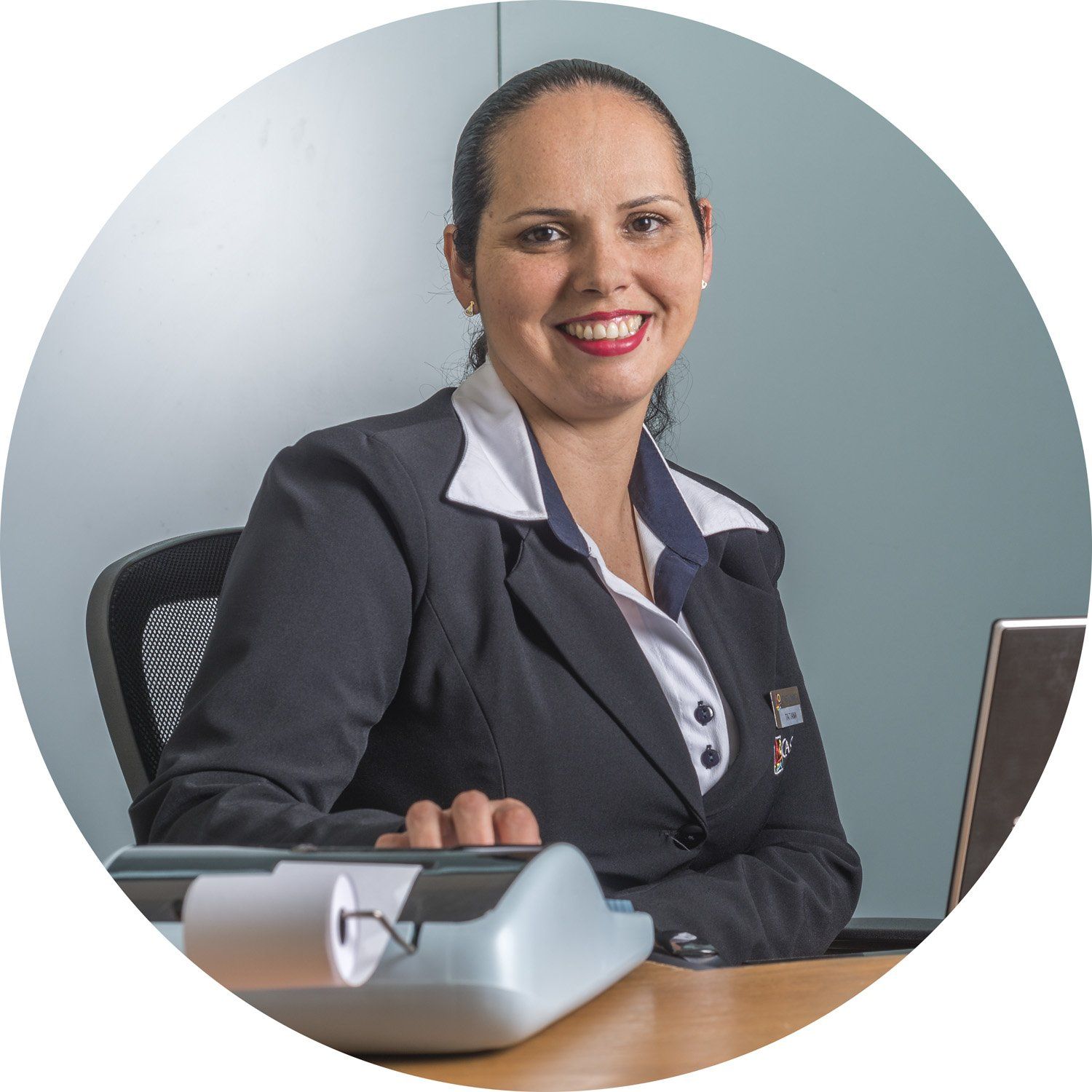 Profissional sorridente sentada à mesa em um escritório, com um laptop e um telefone fixo à sua frente.