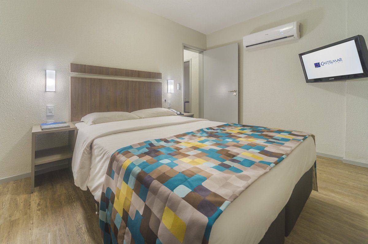 Quarto de hotel com cama, colcha colorida, mesa de cabeceira, TV de parede e ar-condicionado.