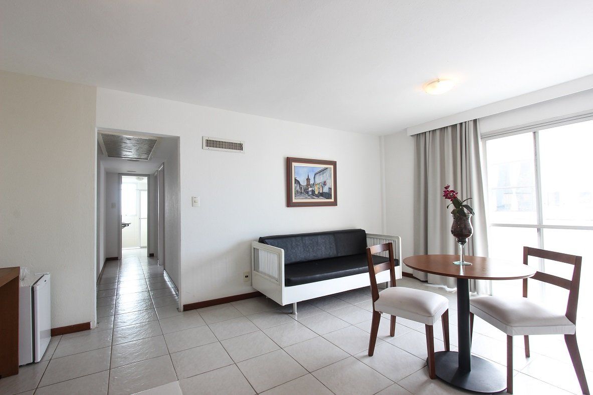 Sala de estar iluminada com sofá preto, mesa de jantar junto à janela e corredor à esquerda.
