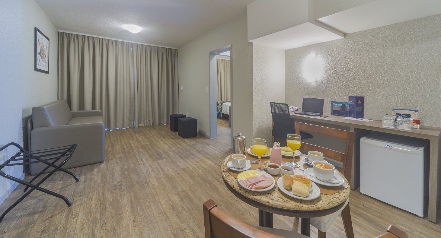 Quarto de hotel pequeno com duas camas de solteiro, um sofá, suporte para TV e mesa de café da manhã posta com alimentos.