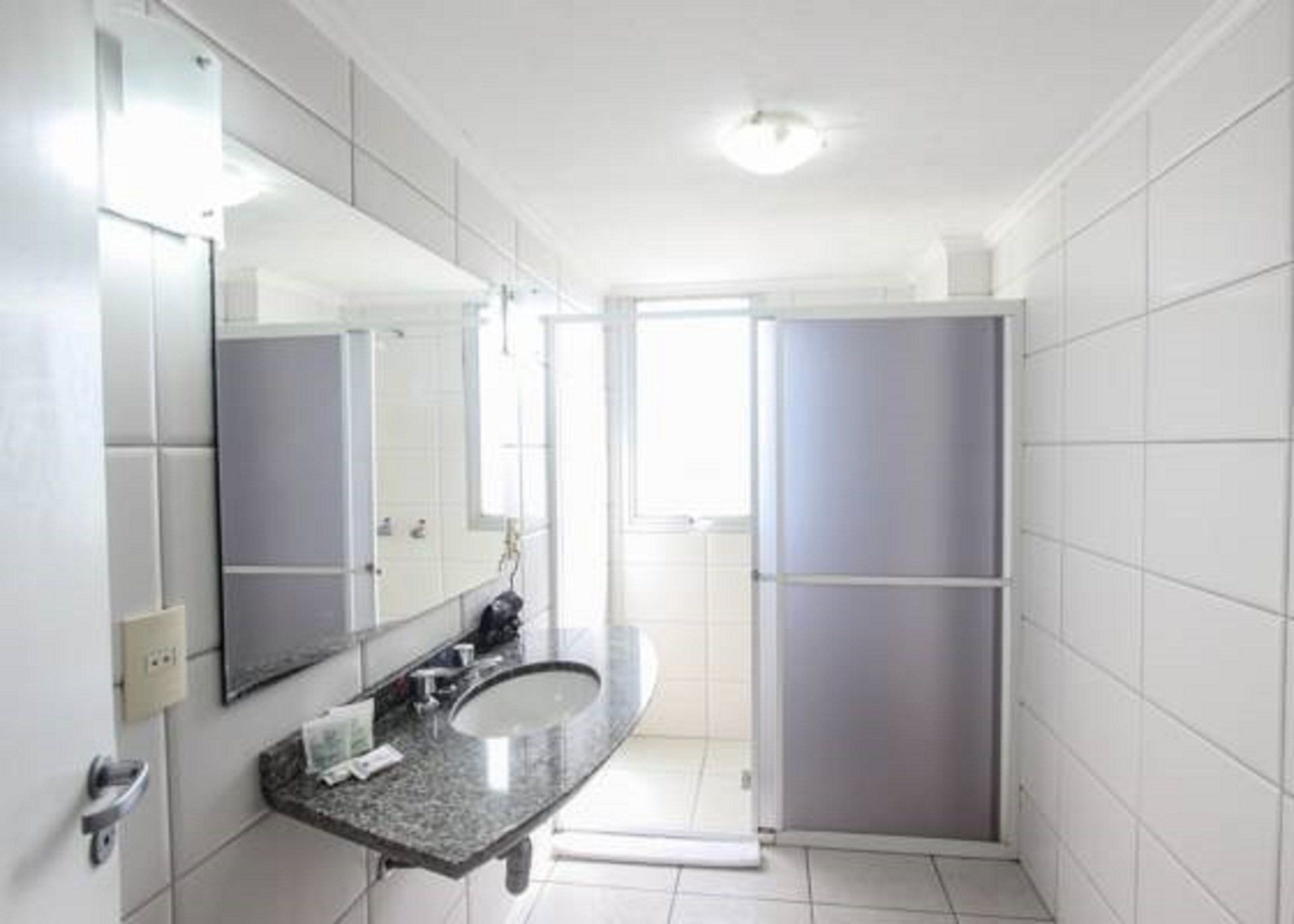 Banheiro com azulejos brancos e luminosos, pia, espelho grande e box para chuveiro.