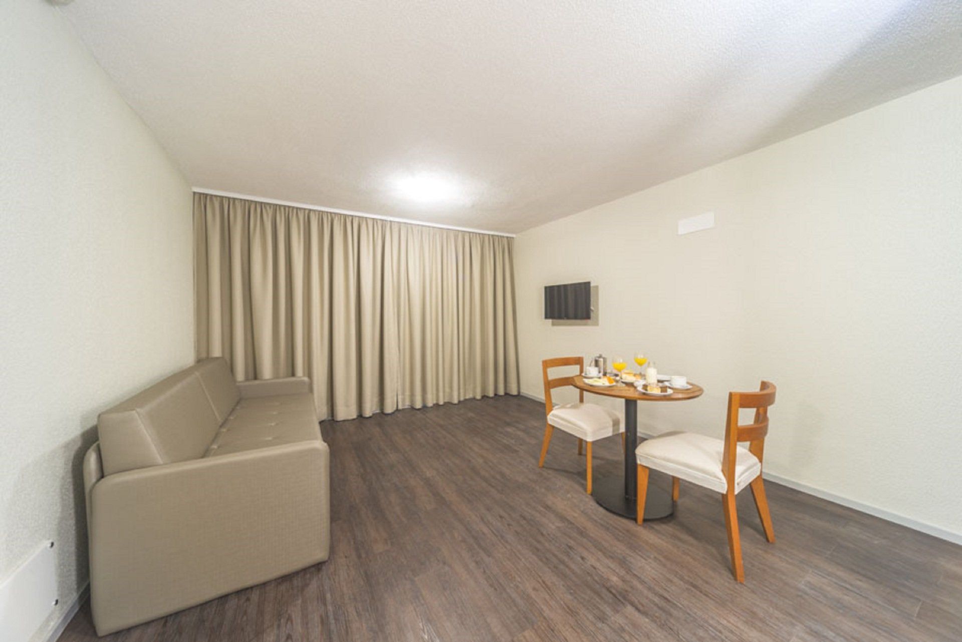 Sala de estar pequena com sofá bege, mesa e cadeiras, TV fixada na parede e cortinas.