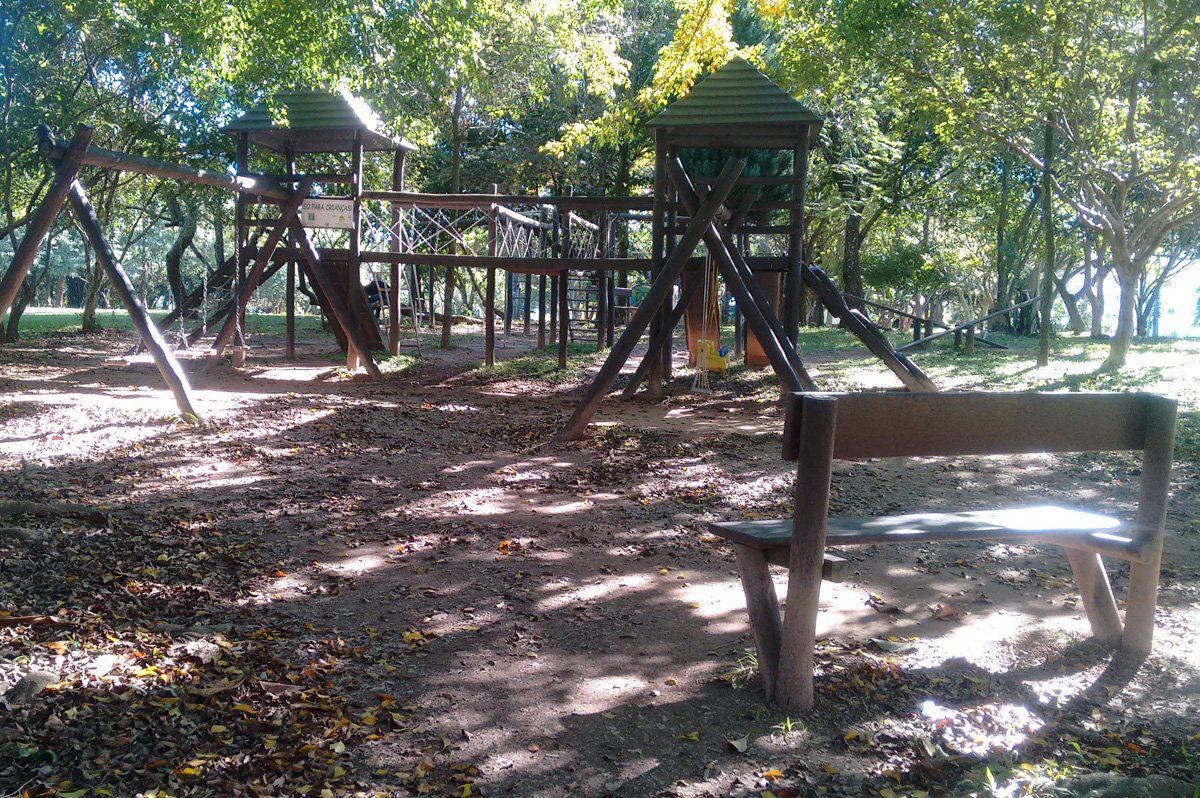 Estrutura de madeira para parque infantil em um parque sombreado, com balanços e um banco em primeiro plano.