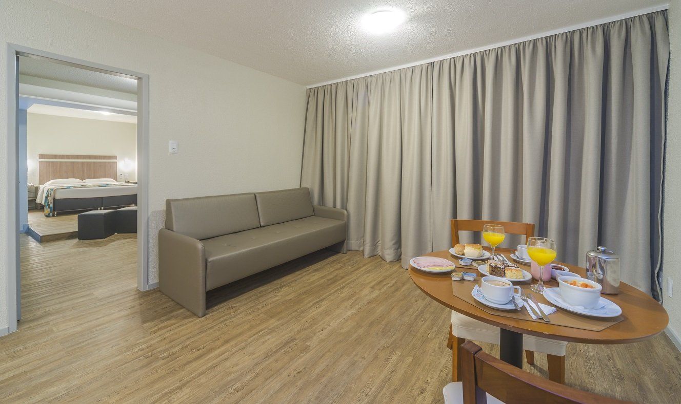 Sala de estar de apartamento pequeno com sofá cinza, mesa de jantar redonda posta para café da manhã e quarto visível através da porta.
