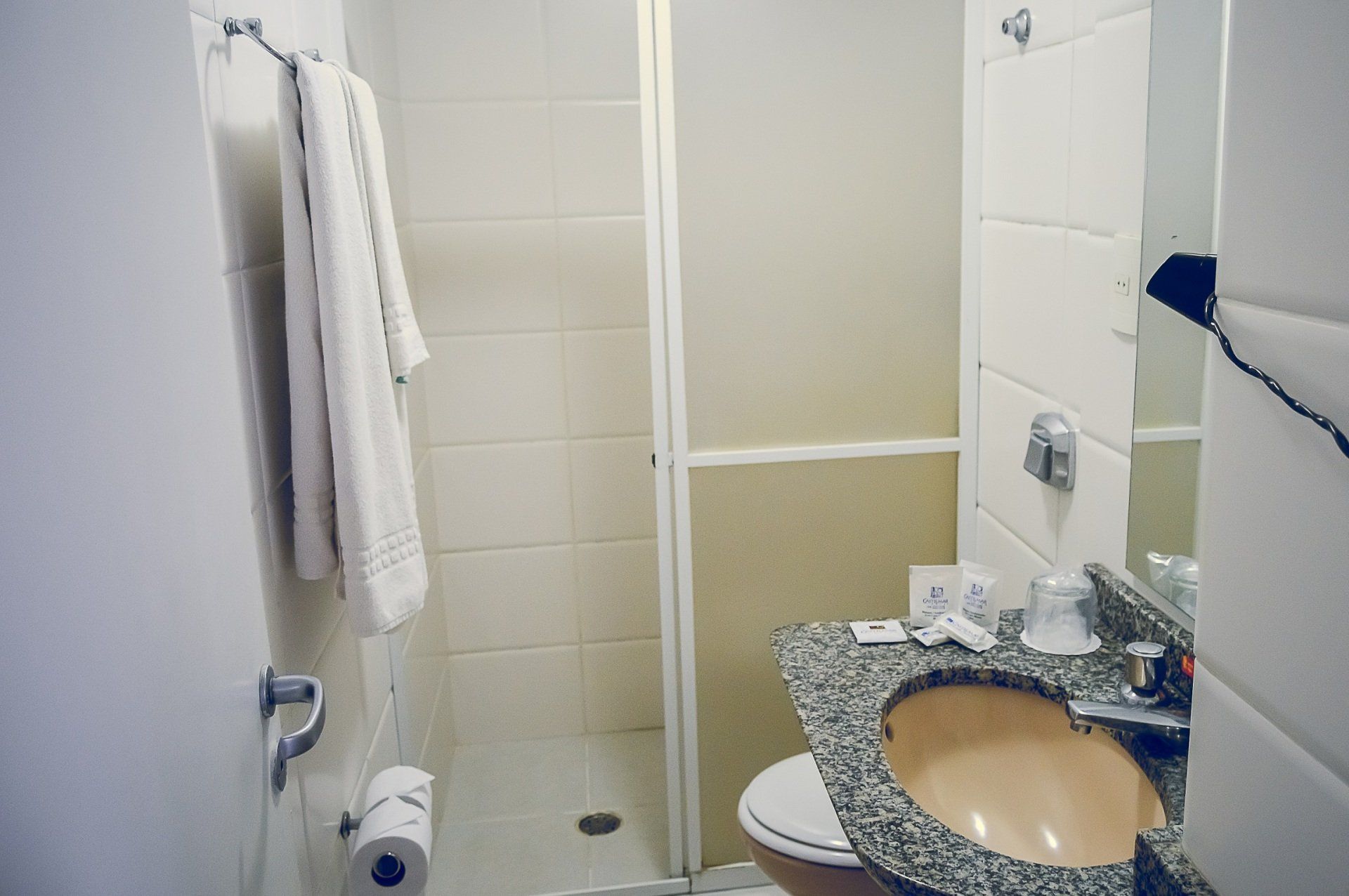 Banheiro pequeno com chuveiro, porta-toalhas e bancada de granito com pia; vaso sanitário visível ao lado de uma divisória de vidro fosco.