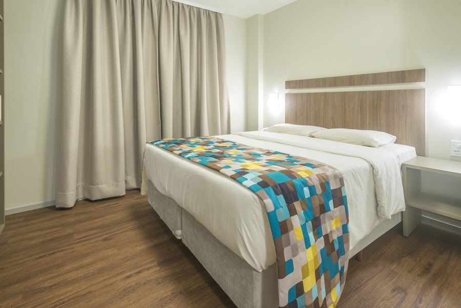 Quarto moderno com cama arrumada, colcha multicolorida e cortinas bege sobre piso de madeira.