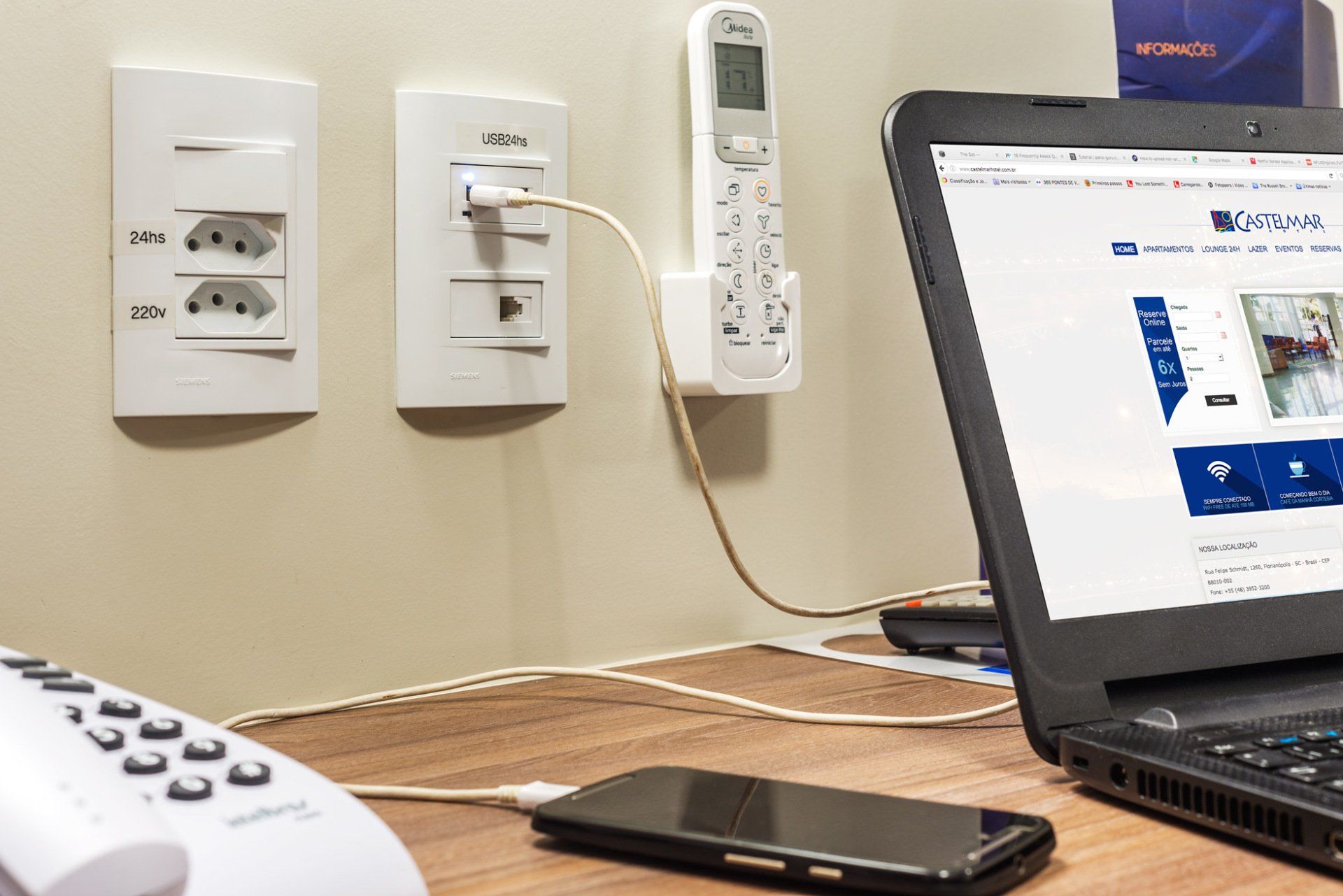 Mesa com laptop, smartphone, telefone fixo e tomadas com carregadores conectados.