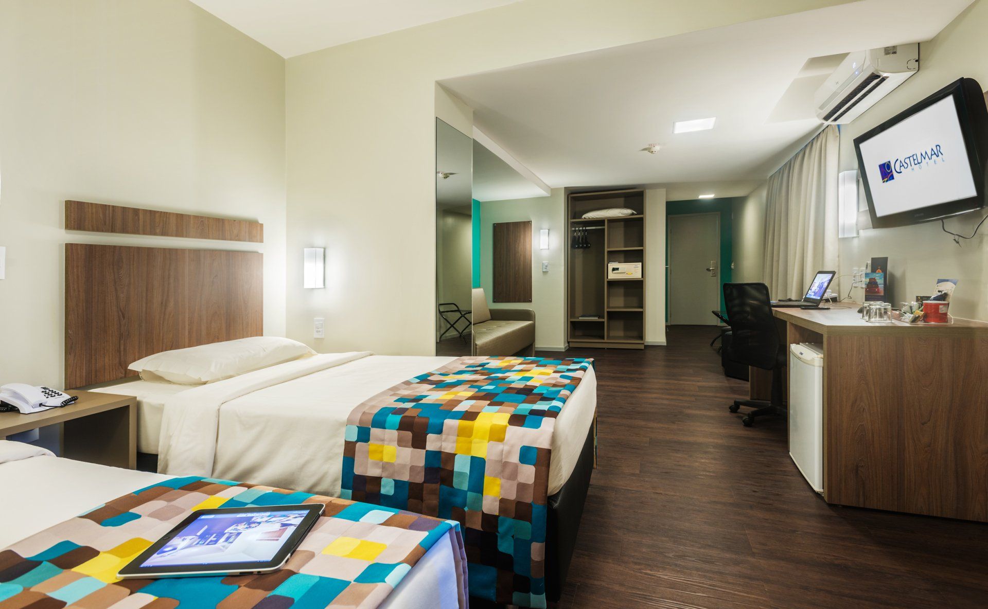 Quarto de hotel moderno com duas camas, móveis de madeira, escrivaninha, TV de parede e piso de madeira escura.