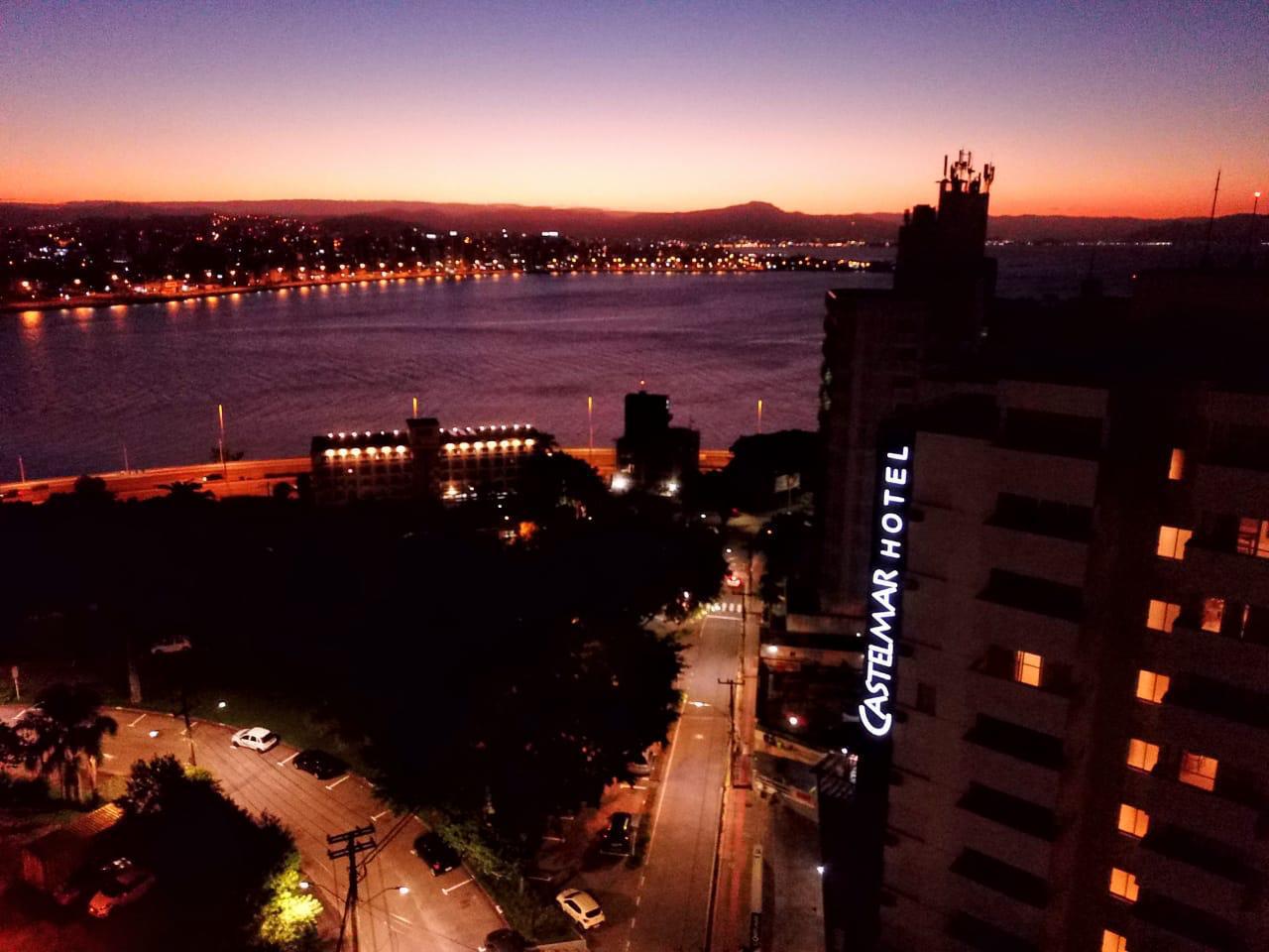 Paisagem urbana ao entardecer sobre um rio, com luzes brilhantes na margem e a placa iluminada do Crown Hotel.