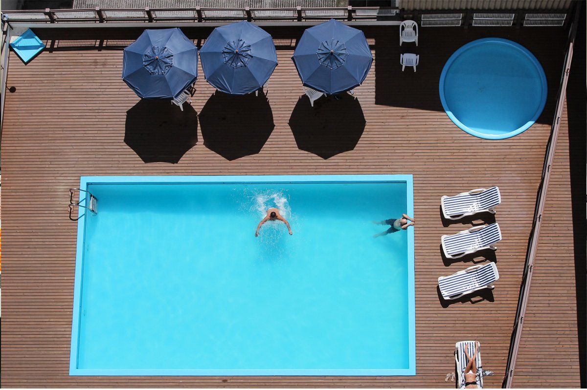 Vista aérea de uma piscina turquesa com nadadores, guarda-sóis azuis e espreguiçadeiras em um deck de madeira.