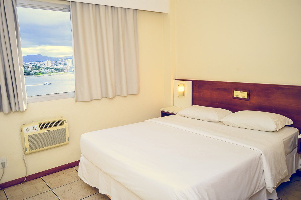 Quarto de hotel pequeno com cama arrumada, janela e ar-condicionado de parede.