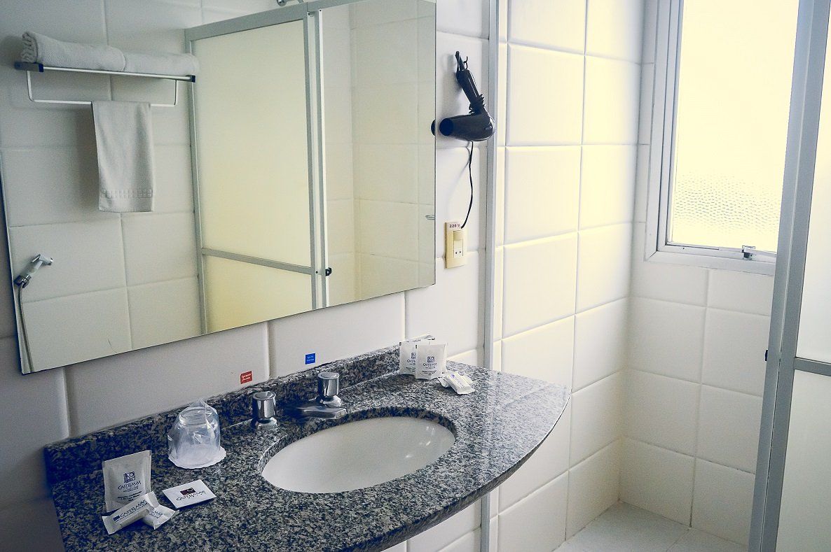 Lavatório com armário espelhado, azulejos brancos e janela fosca ao lado do box do chuveiro.