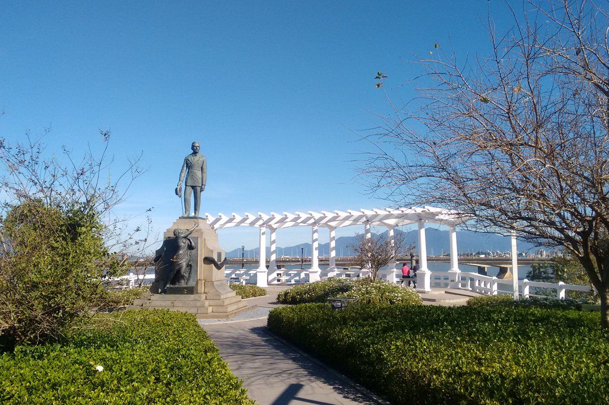 Estátua em um parque ajardinado com uma pérgola branca e céu azul ao fundo.