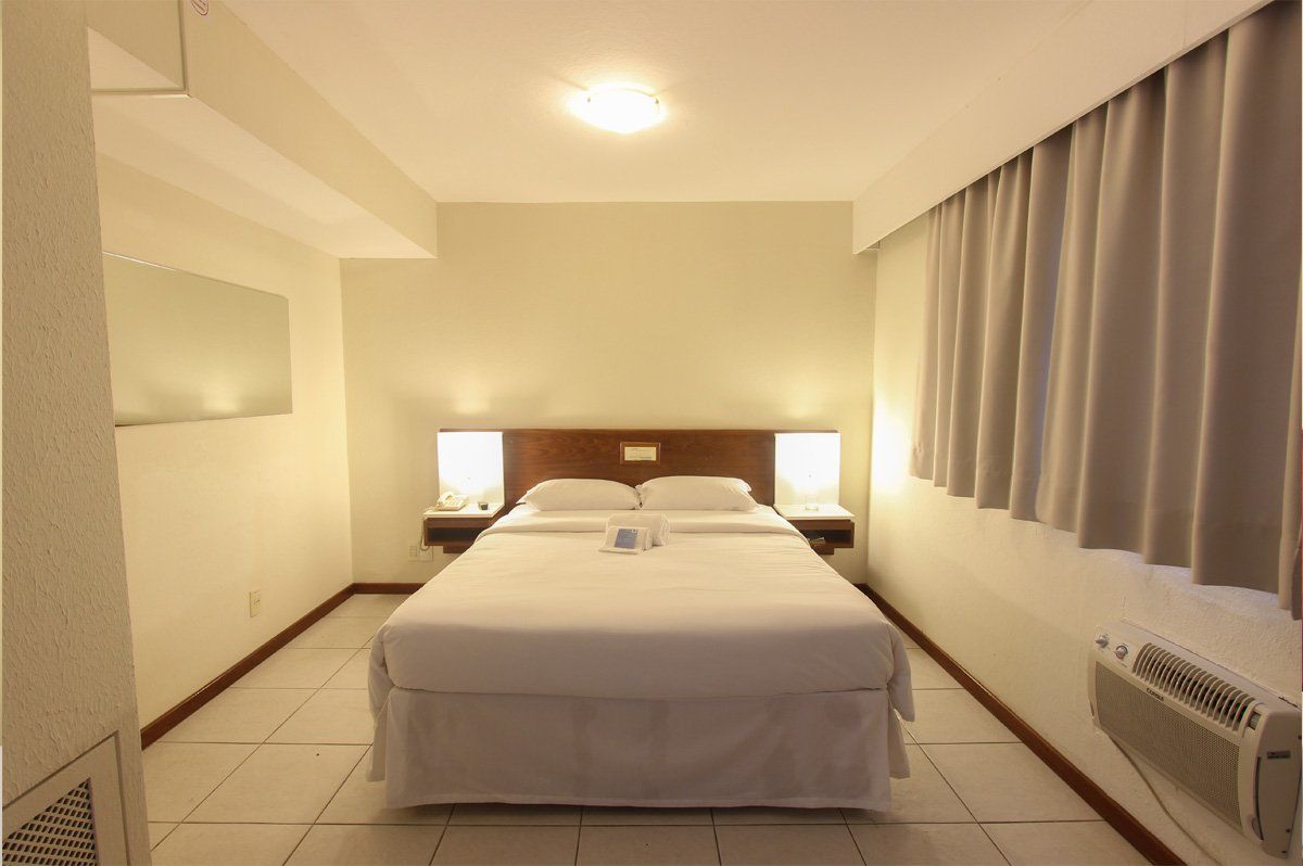 Quarto minimalista em estilo de hotel, com cama arrumada, paredes bege e cortinas na janela.