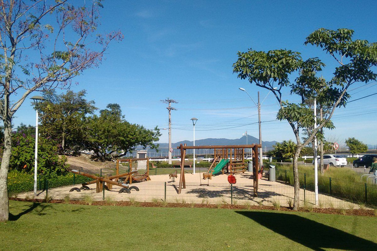 Parque infantil com balanços e estrutura de escalada em um parque ensolarado, cercado por grama e árvores.