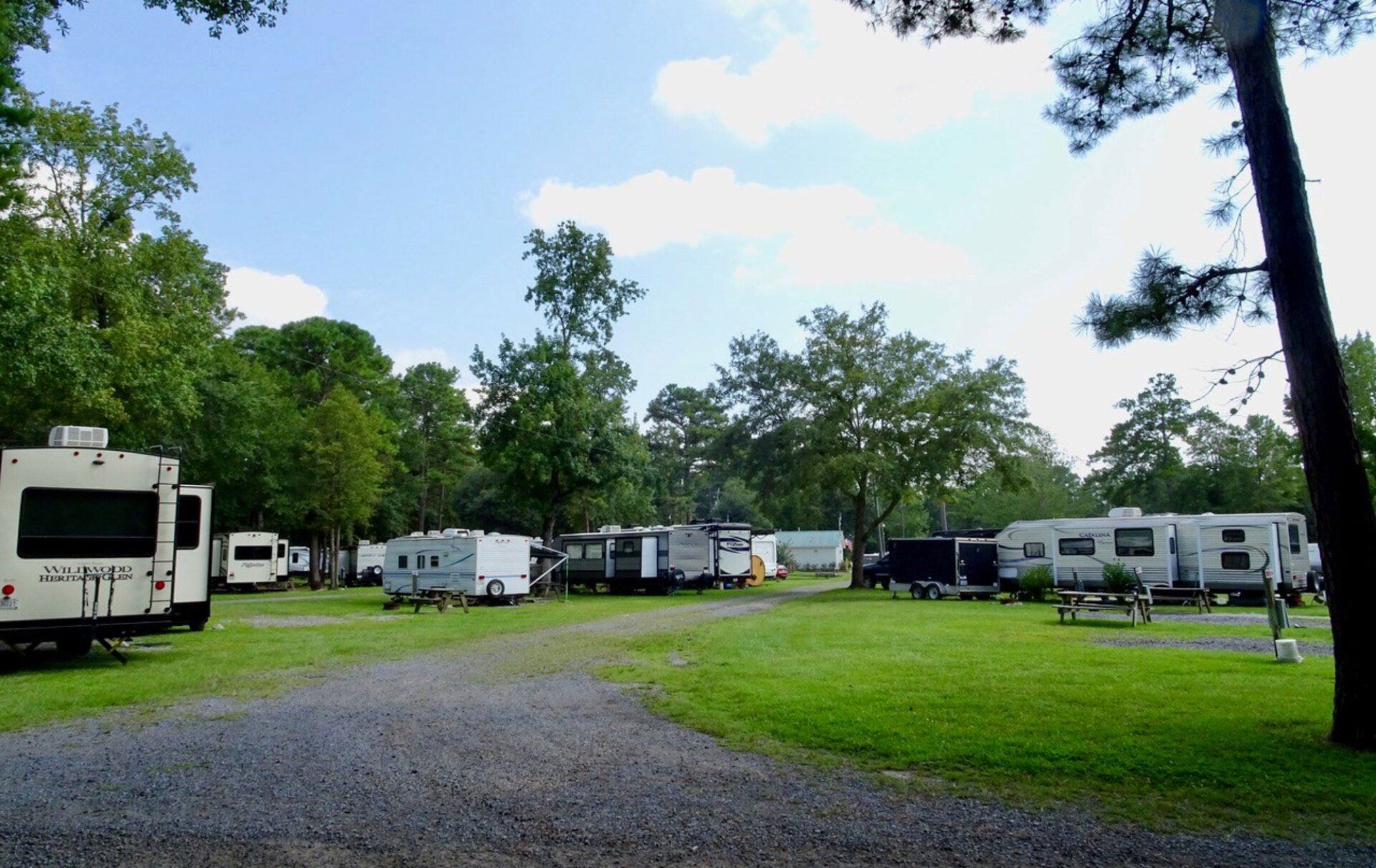 RV park with trailers on grassy sites, gravel road, and trees under a blue sky.
