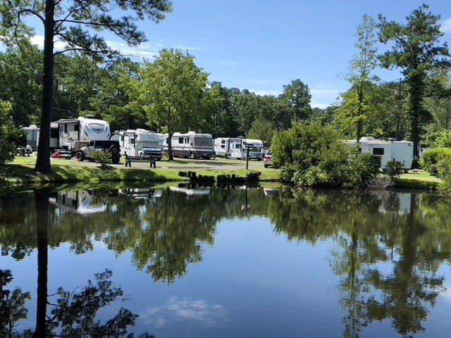 Campground with RVs parked along a calm pond, trees reflect in water on a sunny day.