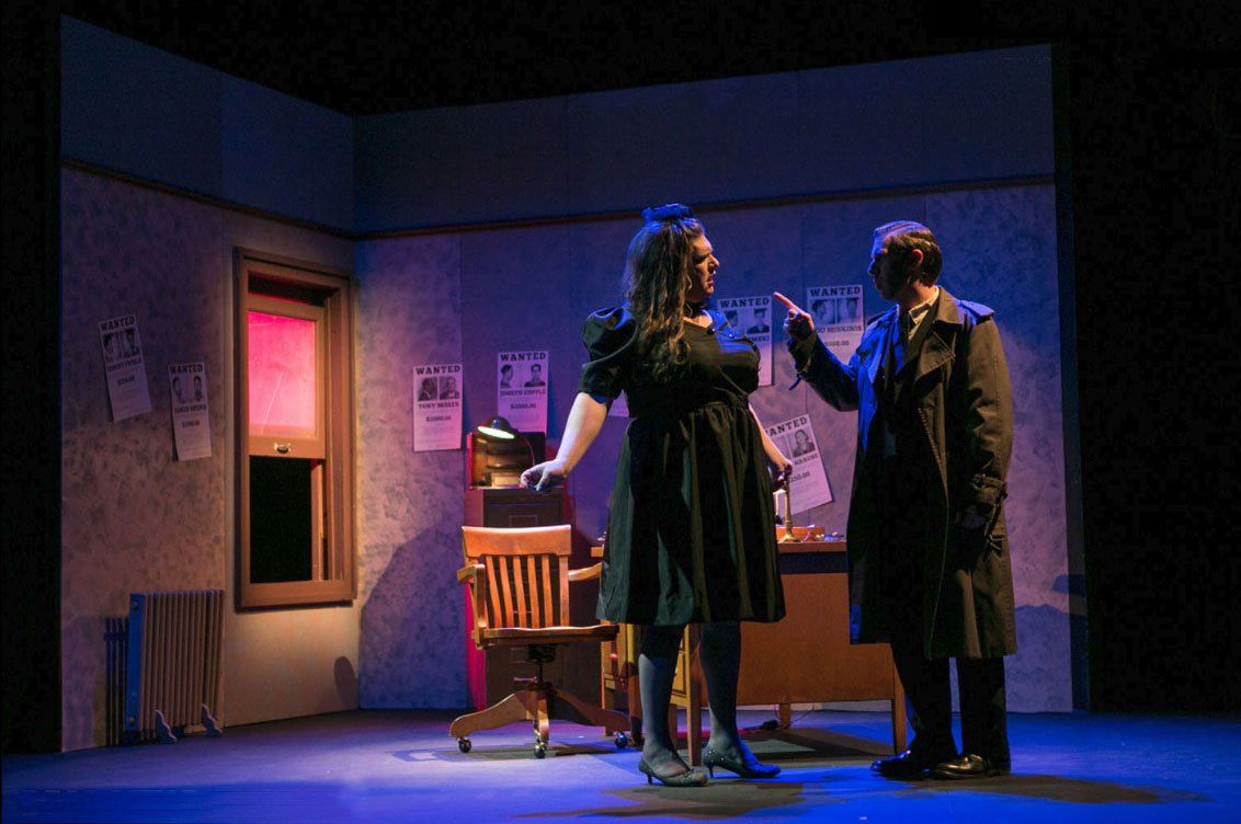 Two people on a stage with a desk, pointing, posters, and a window. Purple and blue lighting.