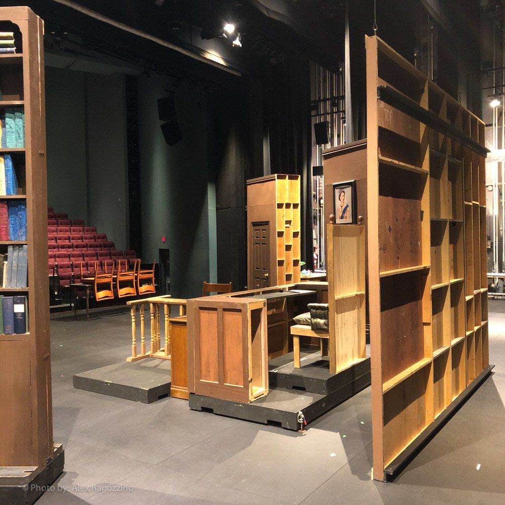 Theater stage set: wood bookshelves, desk, and judge's bench; red auditorium seating in the background.