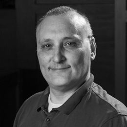 Man with short hair smiles, wearing a polo shirt. Black and white, indoor setting.