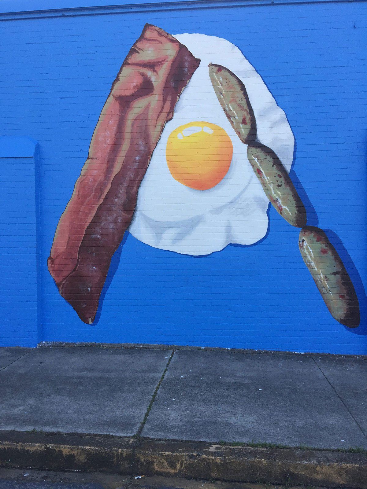A painting of an egg bacon and sausage on a blue wall