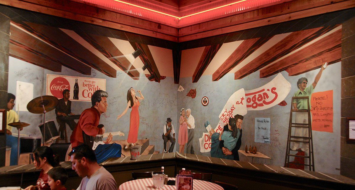A man is sitting at a table in front of a mural of people in a room.