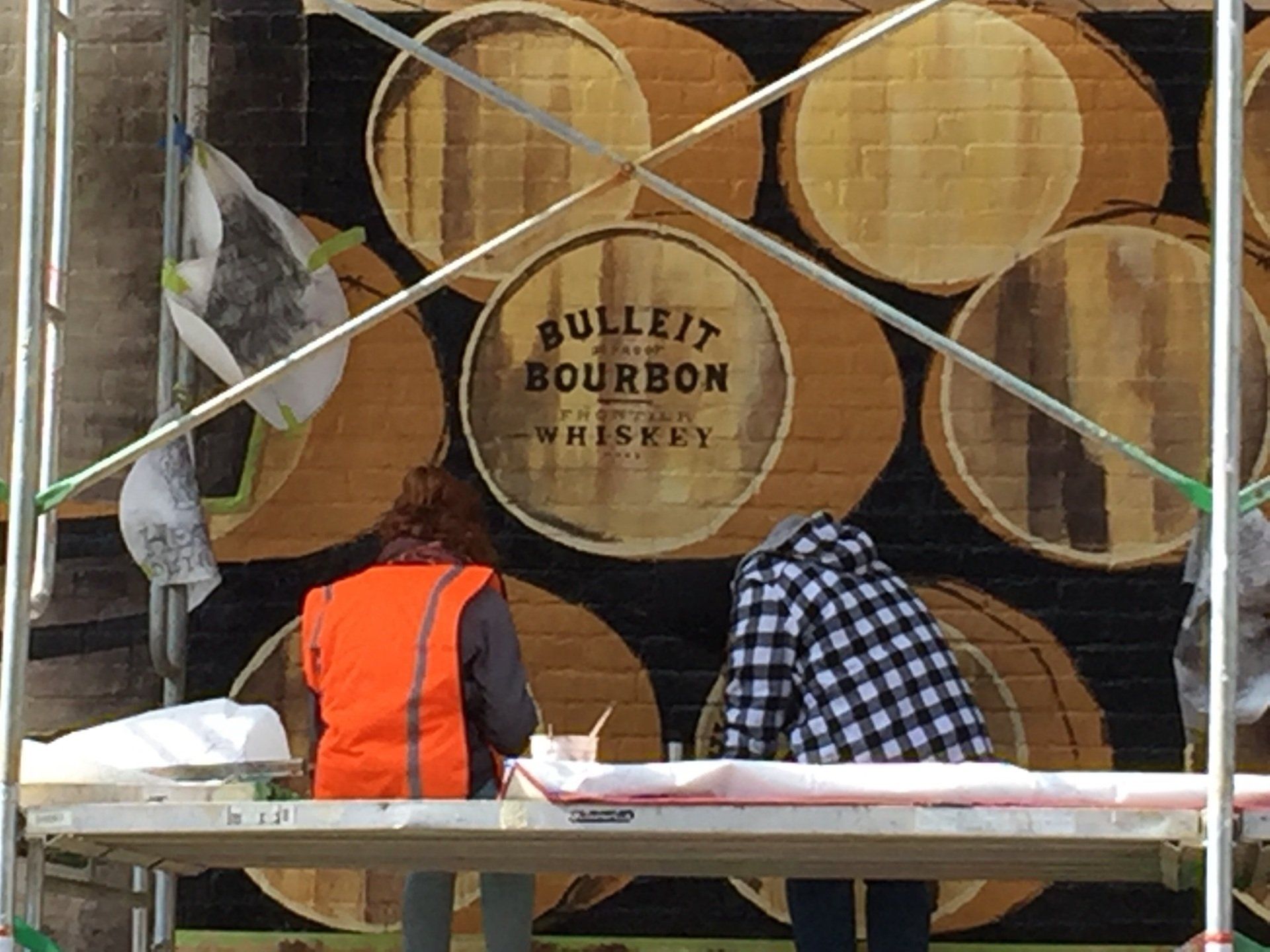 Two people are working on a mural of barrels of bulleit bourbon whiskey.