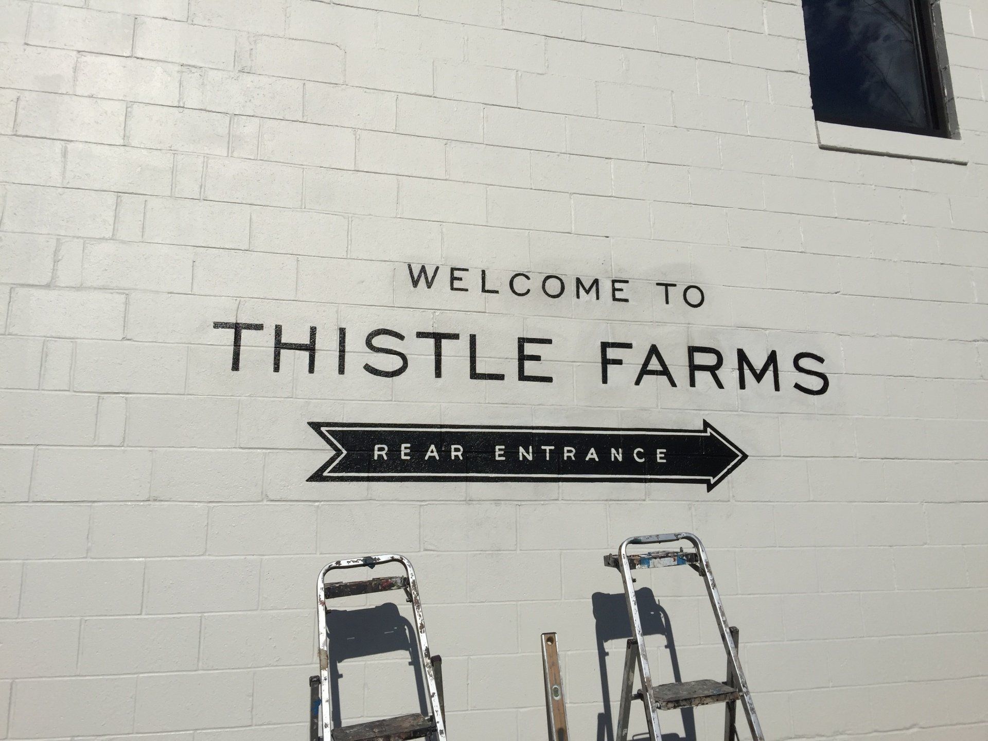 A white brick wall with a sign that says welcome to thistle farms
