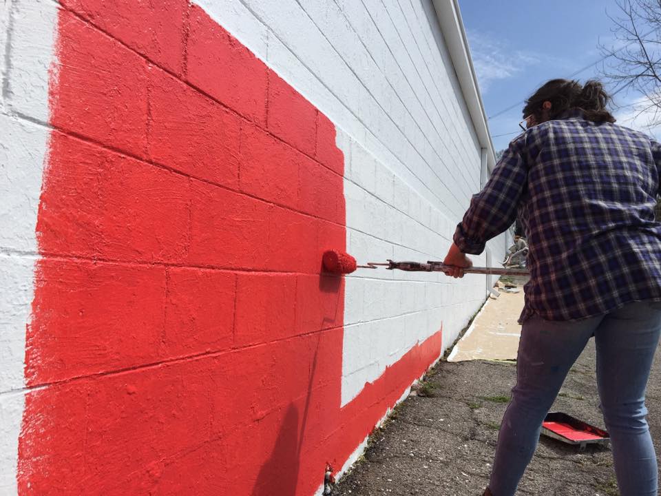 A woman is painting a brick wall red and white with a roller.