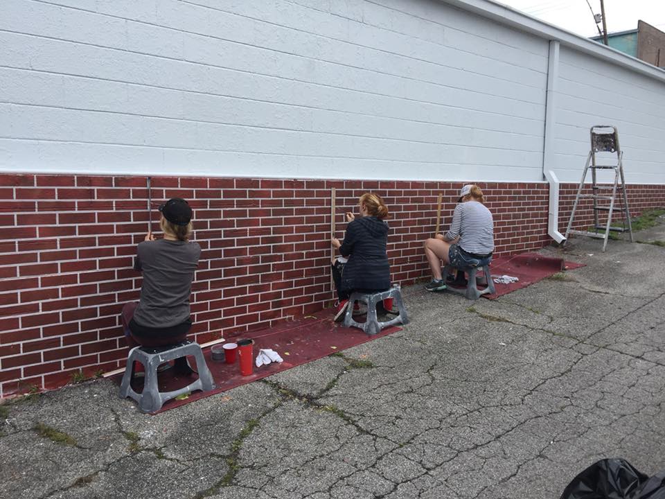 Three people are sitting on stools painting a brick wall.
