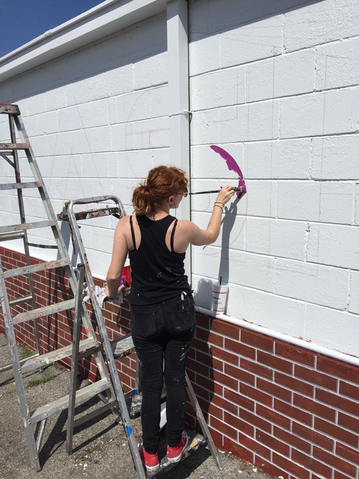 A woman is standing on a ladder painting a brick wall.