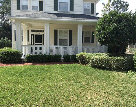 Two-story house with a porch. Beige siding, white trim, green shutters, and a well-manicured lawn with shrubbery.