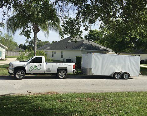 White truck towing a white trailer parked on a street near a house. Landscaping company logo visible on truck.