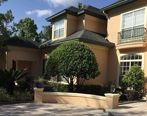 Two-story tan house with black roof, green trees, white windows, and a small wrought-iron balcony.