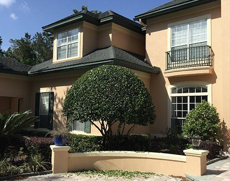 Tan stucco house with dark roof, windows, and small balcony. Large green shrub in front.