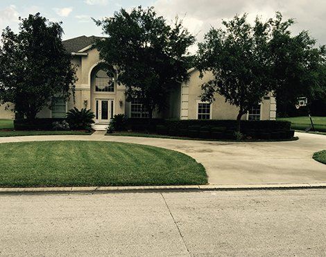 Beige house with arched window and a circular driveway, surrounded by green grass and trees.