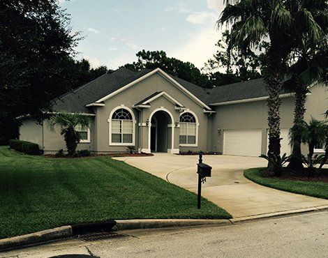 Gray house with a driveway, green lawn, palm trees, and a black mailbox.