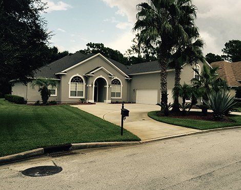 Light gray house with a curved driveway, lawn, and palm trees.
