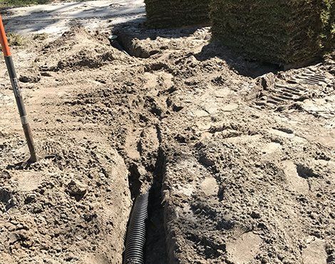 A trench dug in dirt, with black drainage pipe installed. Grass sod in the background.