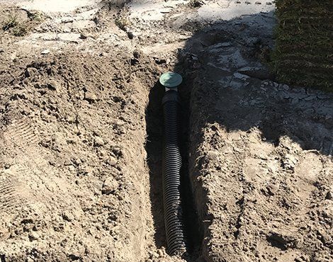 Trench in dirt with black corrugated pipe for irrigation system, capped by green valve, on construction site.