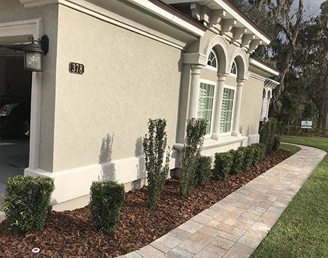 Tan stucco house with white trim, arched windows, and a paved walkway lined with shrubs and mulch.