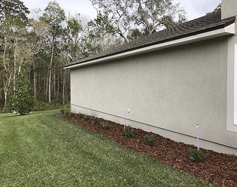 Side view of a light-colored house with a dark roof and a bed of mulch along the foundation; green lawn.
