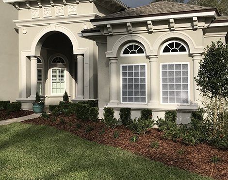 Light gray stucco house with arched entryway, columns, and rectangular windows trimmed in white. Landscaped with mulch and green hedges.
