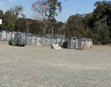 Forklift by stacks of packaged materials on a gravel lot, trees in the background.