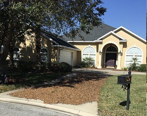 Beige house with arched windows and a dark roof. Driveway has mulch. Green lawn and a mailbox.