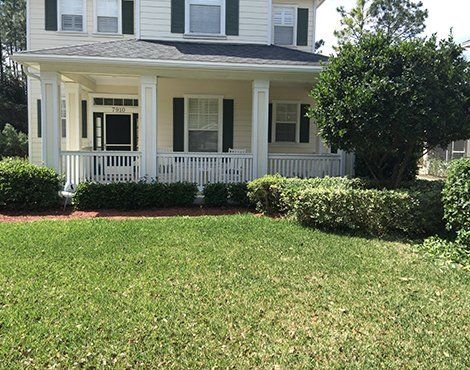 A two-story house with a porch and green lawn. White siding, black shutters, green bushes.