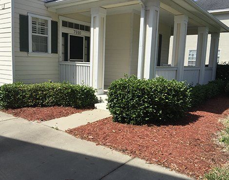 A house with a white porch, trimmed bushes, and red mulch in front.