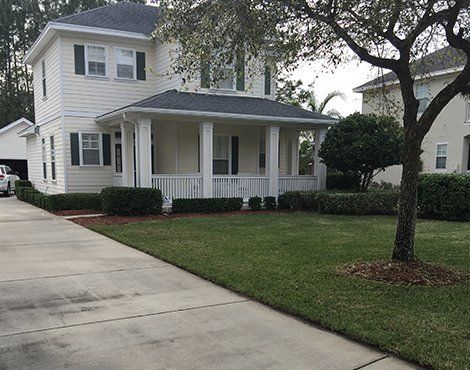 Two-story white house with green shutters, front porch, and manicured lawn. Driveway in foreground, tree on right.