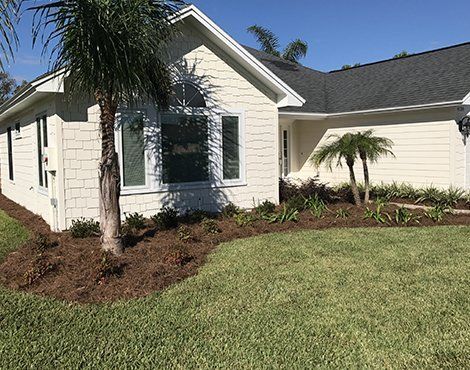 Cream-colored house with dark roof. Palm trees and landscaping in front. Green grass. Blue sky.